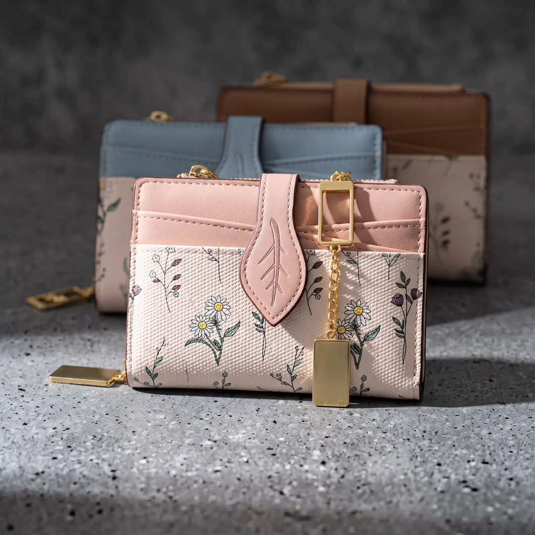 Compact bi-fold wallet featuring an off-white floral print with pink accents and a unique leaf-shaped tab closure, accented with gold-tone hardware and a dangling charm.