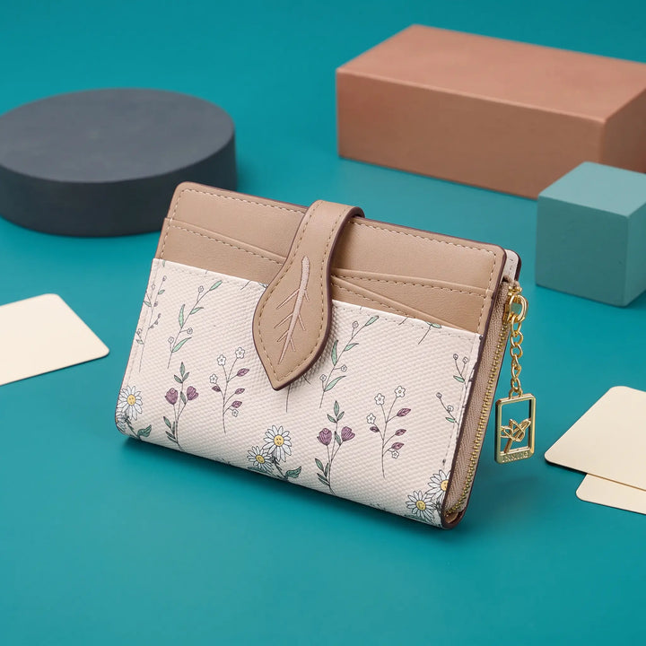 Compact bi-fold wallet in off-white with a delicate multi-colored floral print, featuring a tan leaf-shaped tab closure and a gold-tone zippered coin pocket on the back.