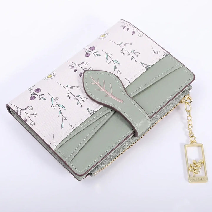 Compact bi-fold wallet in sage green and off-white with a delicate floral print, featuring a leaf-shaped tab closure and gold-tone hardware. This chic accessory offers convenient organization for cards and coins.