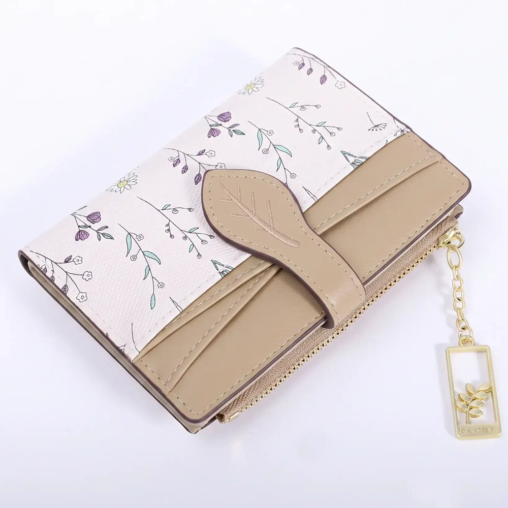 Compact bi-fold wallet in off-white with a delicate multi-colored floral print, featuring a contrasting tan flap, leaf-shaped closure, and a gold-tone zipper pocket with a charm.