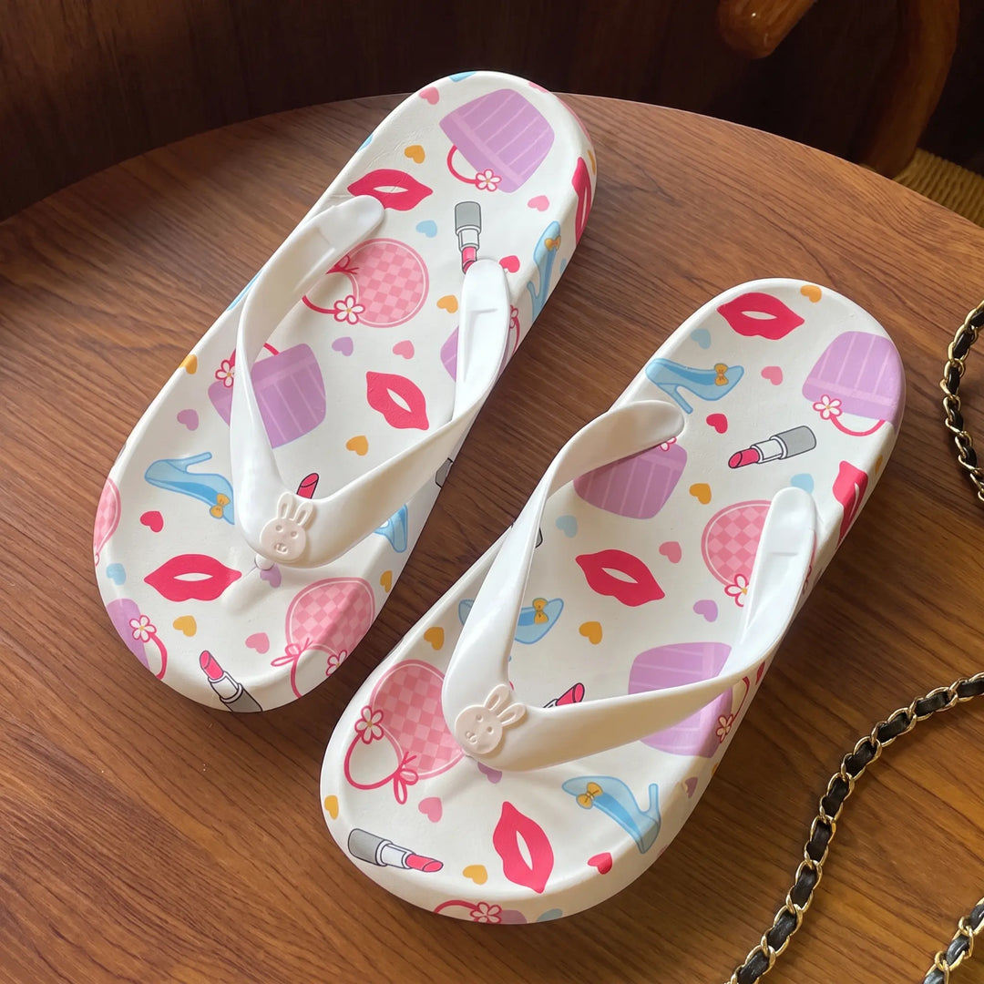 A pair of white flip-flop sandals adorned with a playful print of lips, handbags, lipstick, and hearts, featuring a small bunny charm on the toe post, laid on a wooden surface.