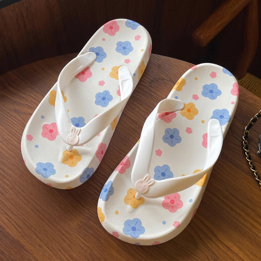 Pair of white platform flip-flops adorned with pastel floral prints and a cute pink bunny charm on the toe post, displayed on a wooden surface.