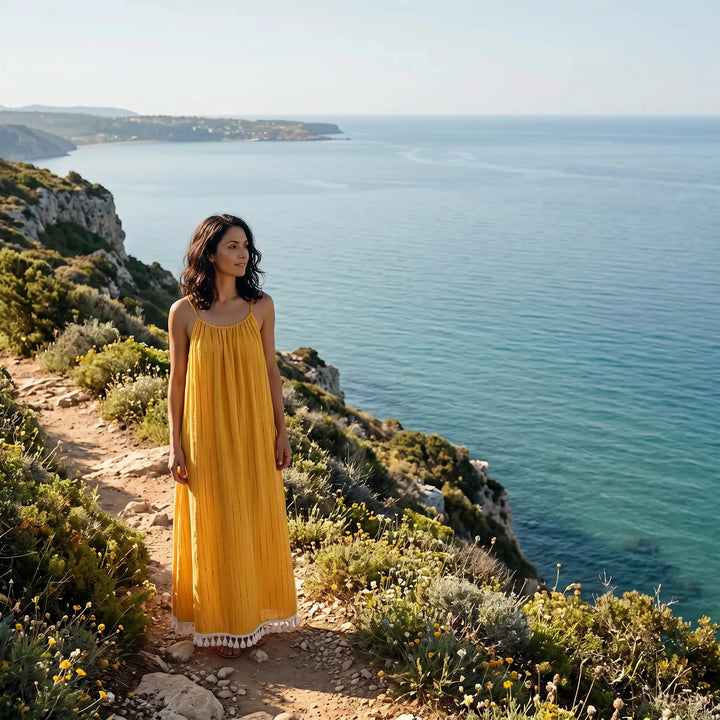 Woman in a golden yellow bohemian maxi dress with fine linear embroidery and a white tassel hem, standing on a scenic coastal path overlooking the blue ocean.