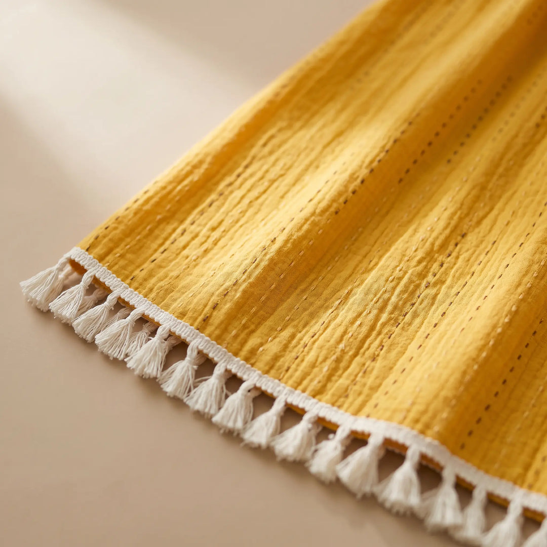Close-up of a golden yellow maxi dress hem featuring fine linear tonal embroidery and a delicate white tassel trim.