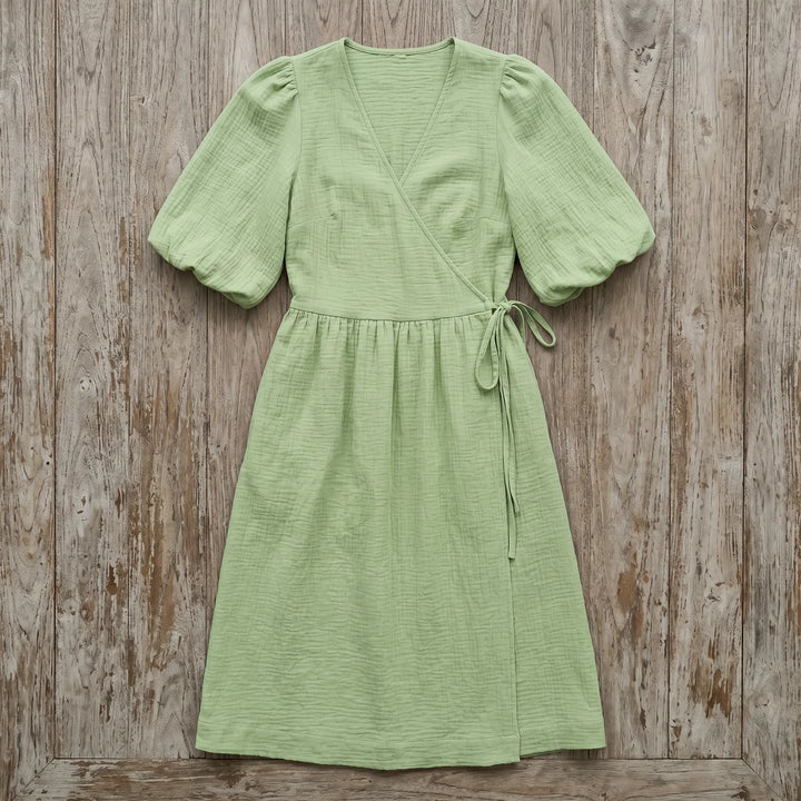 Light green crinkled cotton gauze midi wrap dress with short puff sleeves and a tie waist, presented as a flat lay on a rustic wooden background.