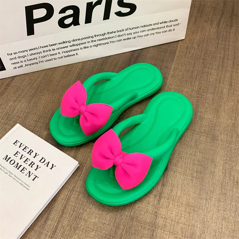 Green flip-flops for women featuring bright pink bow embellishments with a charming teddy bear charm, displayed on a wooden surface next to a book.