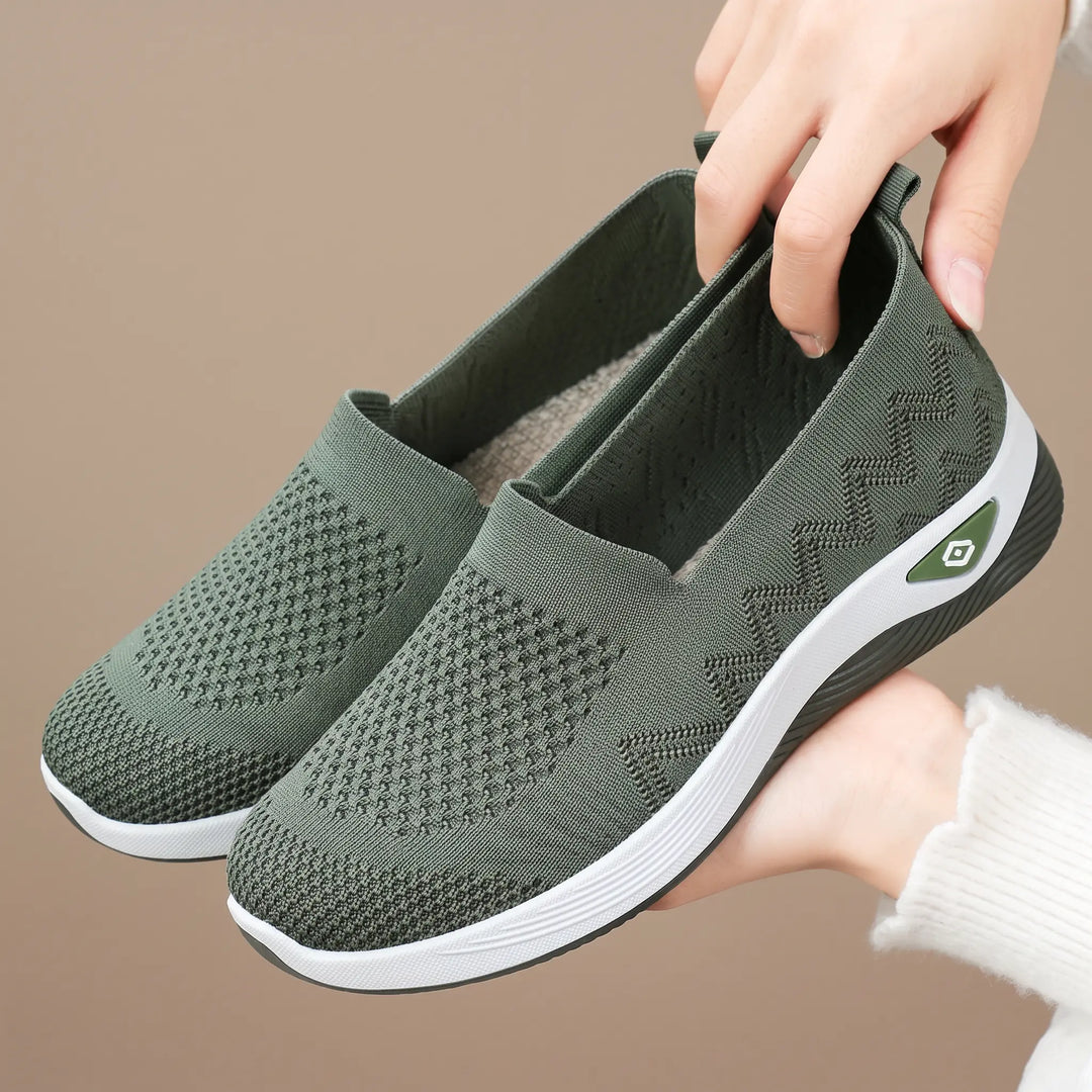 Green engineered knit slip-on sneakers with diamond-patterned vamp and chevron heel, held by hands against a soft background.