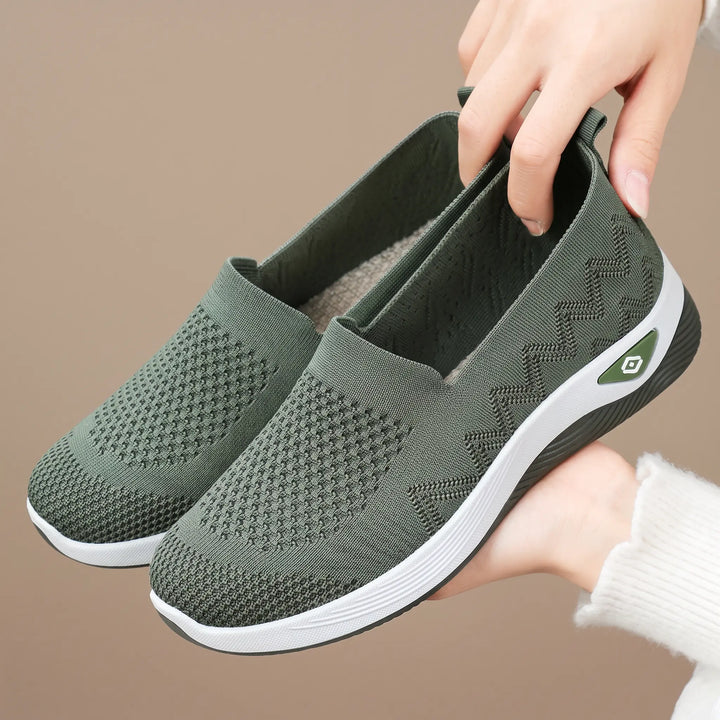 Green engineered knit slip-on sneakers with diamond-patterned vamp and chevron heel, held by hands against a soft background.