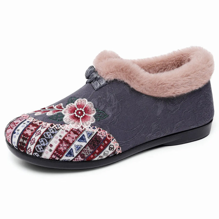 Grey woven mule slipper with intricate colorful floral embroidery and geometric band, featuring plush faux-fur lining and a decorative bow accent, displayed on a white background.