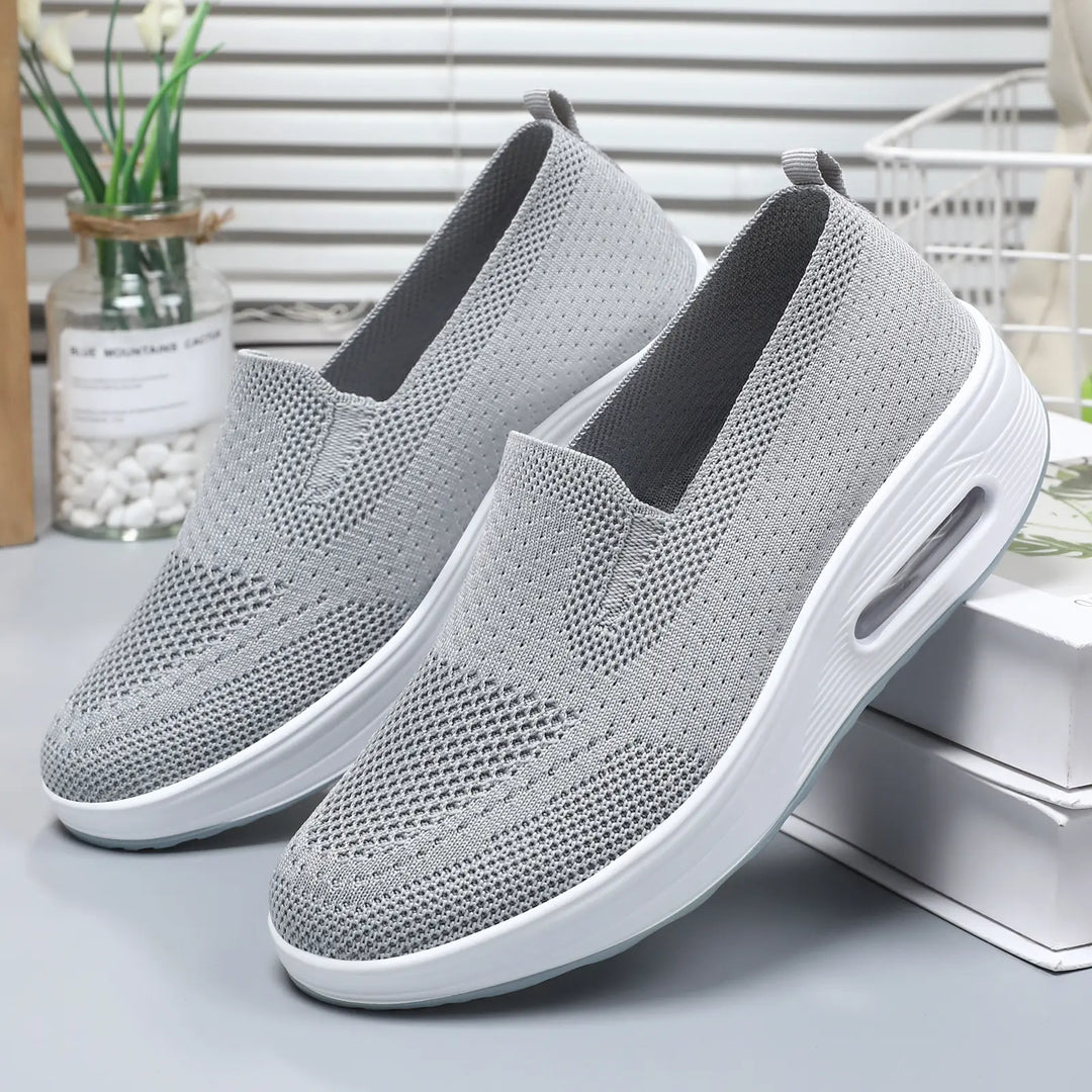 A pair of grey breathable knit slip-on sneakers with white platform soles and a visible air cushion sole detail, shown from a front three-quarter angle on a neutral studio surface.