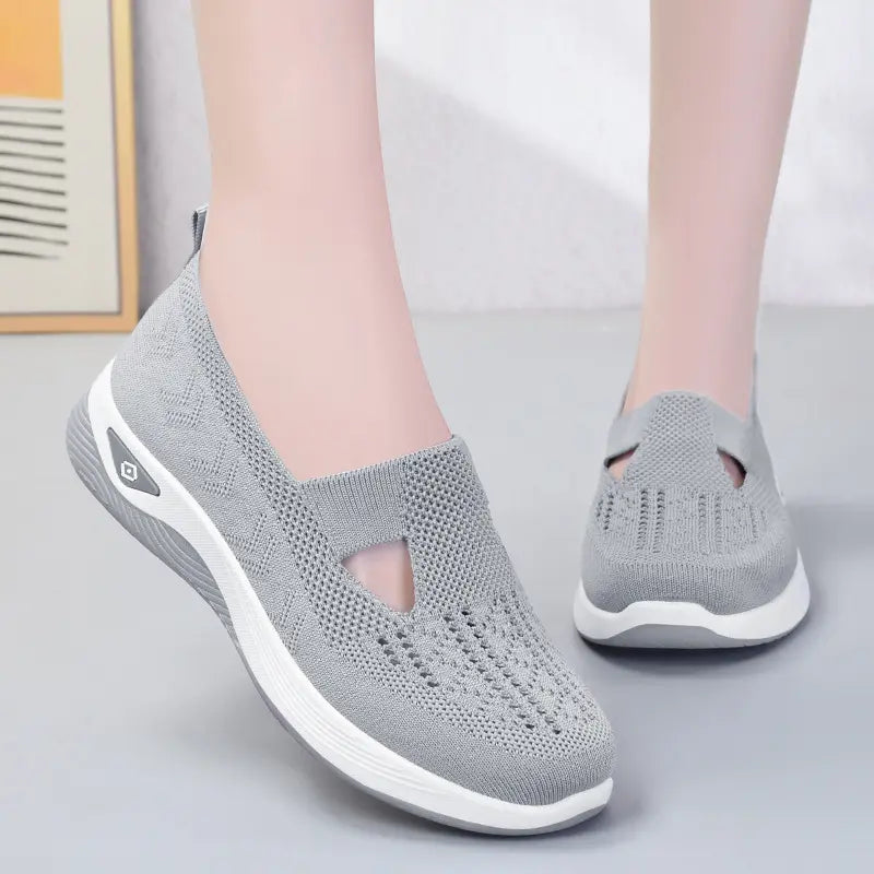 A pair of grey breathable knit slip-on sneakers worn on feet, showcasing their flexible upper and cushioned white sole with a subtle geometric accent.