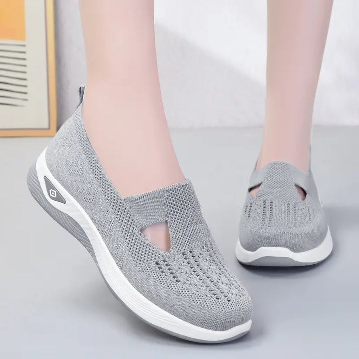 A pair of grey breathable knit slip-on sneakers worn on feet, showcasing their flexible upper and cushioned white sole with a subtle geometric accent.