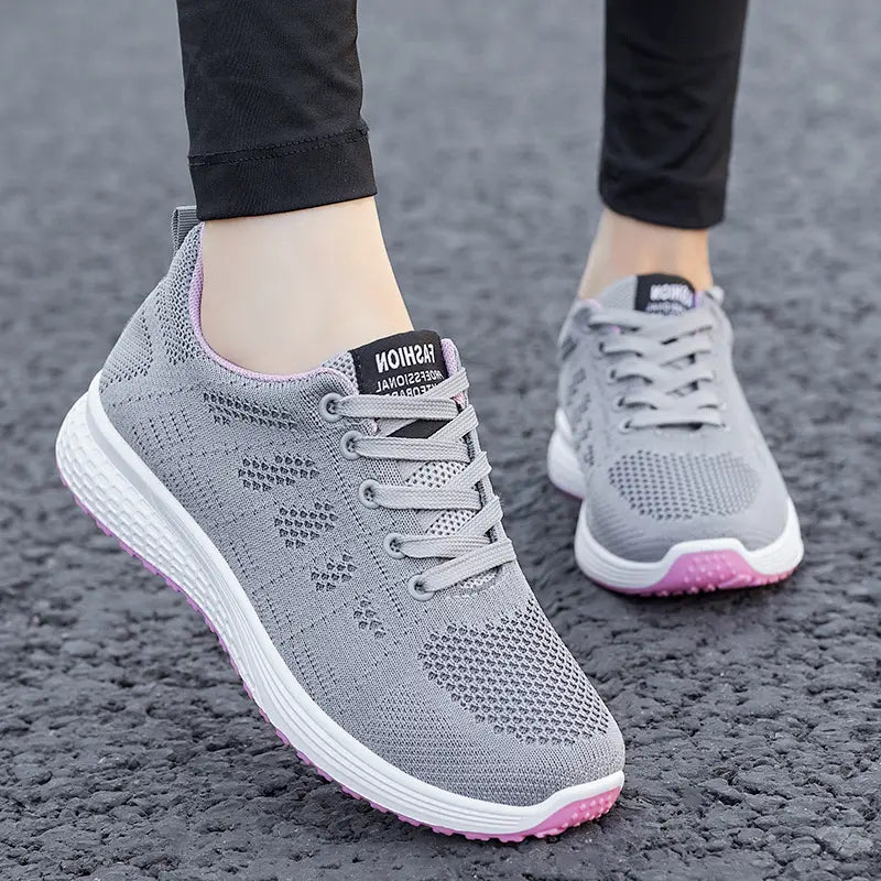 Grey knit low-top athletic sneakers featuring a delicate pink geometric pattern, plush pink lining, white midsole with a textured wave pattern, and a vibrant pink rubber outsole, shown from a three-quarter view on a paved surface.