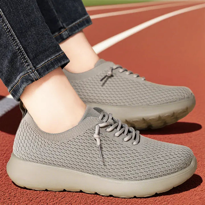 Grey engineered knit trainers worn by a person on a red running track, showcasing their breathable, sock-like fit and comfortable cushioned sole.