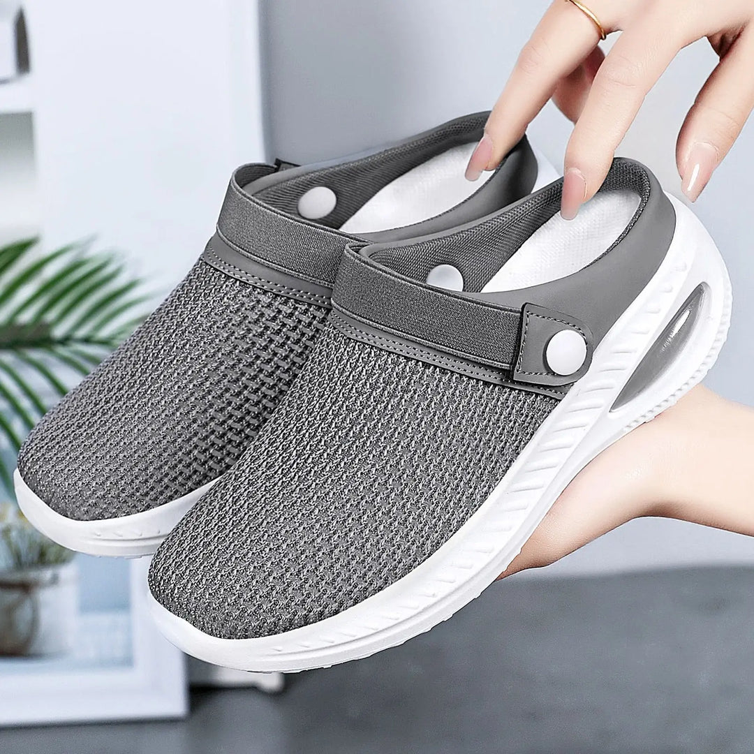 Grey breathable mesh slip-on clogs with a white air cushion sole and adjustable strap, shown held by hands against a soft-focus background.