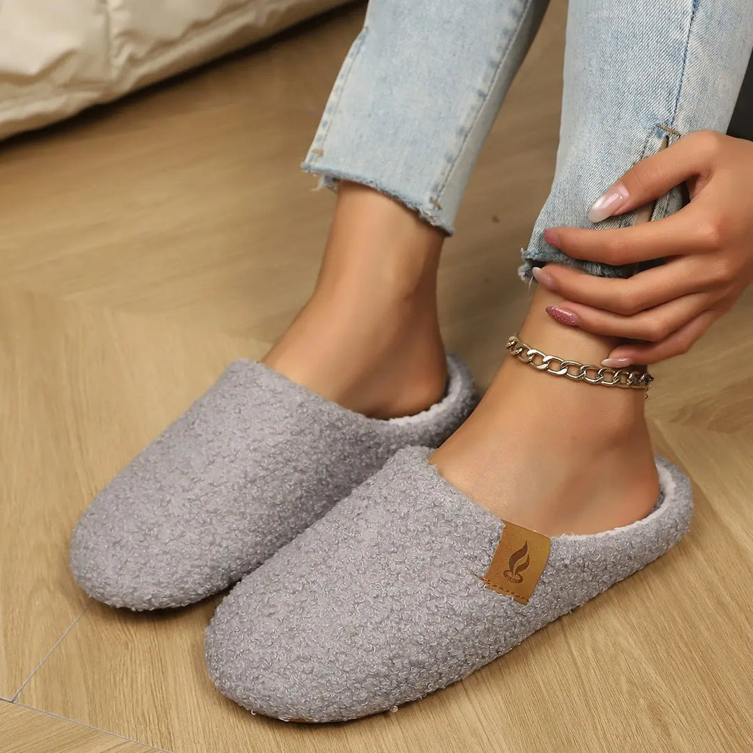 A pair of grey plush clog slippers worn by a person, showcasing their soft texture and white lining, styled with denim jeans and an ankle bracelet on a wooden floor.