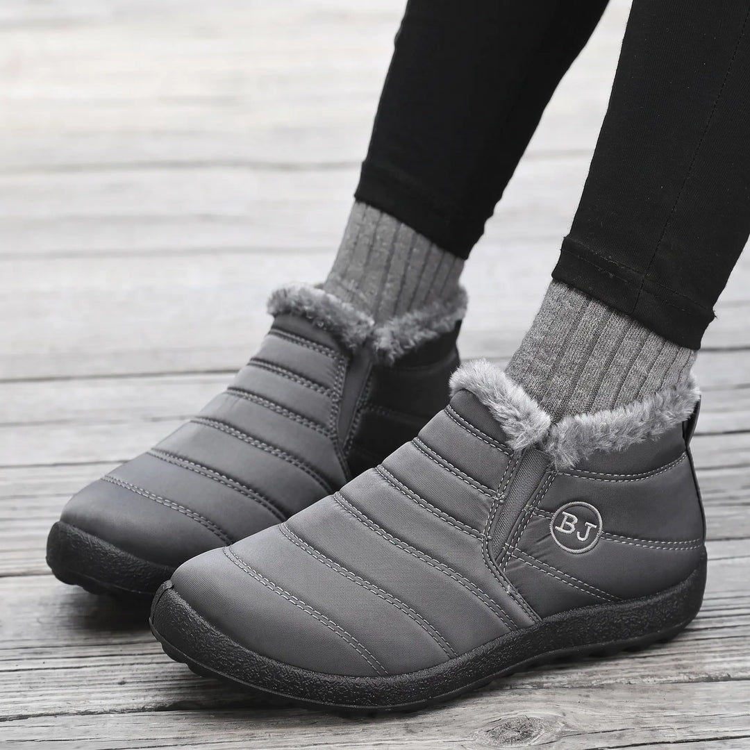 Grey quilted waterproof ankle boots with fluffy faux fur trim and side gore panels, worn with grey socks and black leggings on a wooden floor.