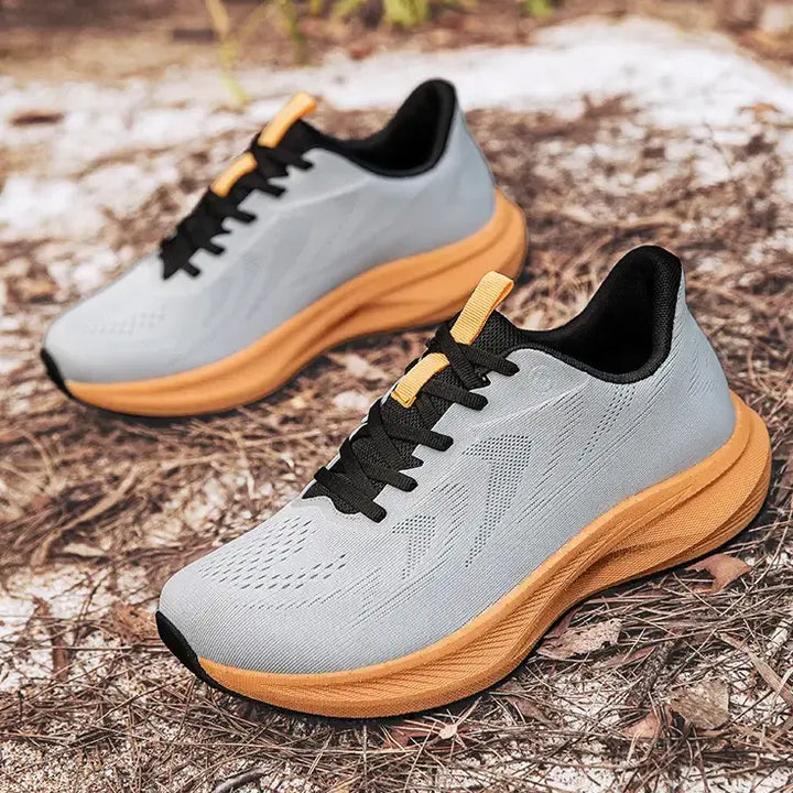A pair of light grey wide-fit athletic trainers with vibrant orange soles and black laces, shown resting on a textured outdoor ground surface.