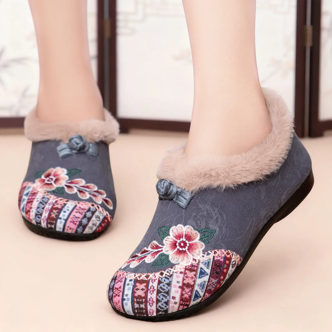 Grey embroidered mule slippers for women, featuring a plush faux-fur lining, colorful floral embroidery on the vamp, and a decorative bow accent. These slip-on shoes offer comfort and style.