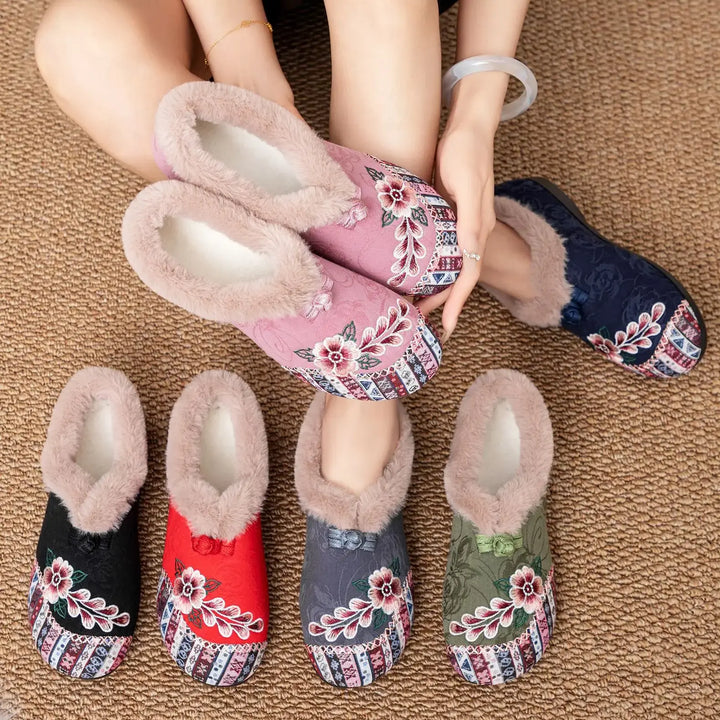 A collection of Grivan embroidered mule slippers in various colors including black, red, grey, green, pink, and navy, featuring plush faux-fur lining and intricate floral embroidery. A pair of pink slippers are held by a person wearing a bracelet and ring.