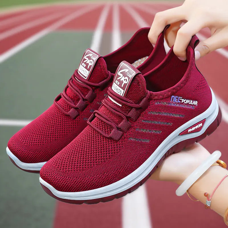 A pair of deep red Gryphos breathable mesh sneakers with white midsoles, shown held by hands against a blurred sports track background. Features include elastic laces, heel pull-tabs, and subtle branding text.