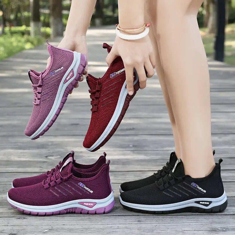 Two pairs of Gryphos breathable mesh sneakers, one held in deep purple and burgundy red, and another pair worn in deep purple and black, shown outdoors on a wooden walkway.