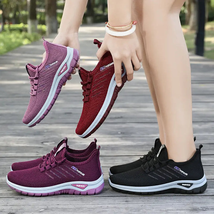 Two pairs of Gryphos breathable mesh sneakers, one held in deep purple and burgundy red, and another pair worn in deep purple and black, shown outdoors on a wooden walkway.