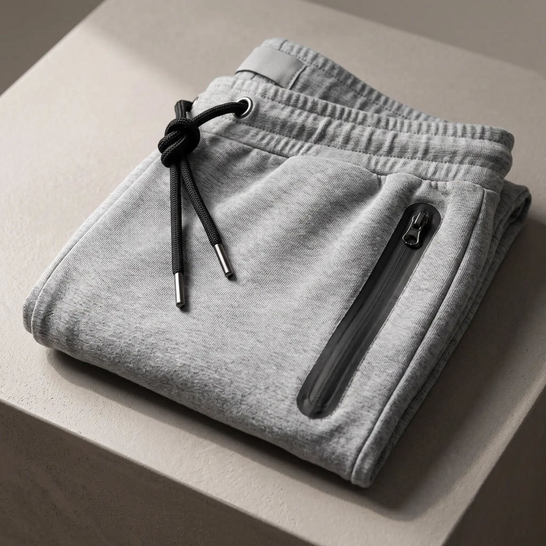 Close-up of heathered grey performance knit joggers, showcasing the subtle fabric texture, flat elasticated waistband with drawstring, and a functional zippered side pocket with a black pull tab.