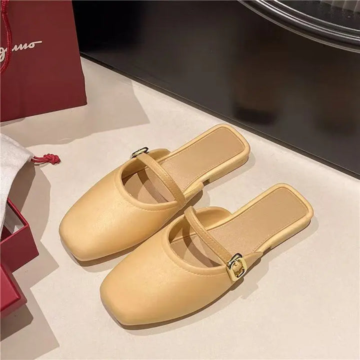 A pair of soft ivory faux leather square-toe mules with a slender strap and polished gold-tone buckle, shown in a three-quarter view on a studio surface with packaging elements.