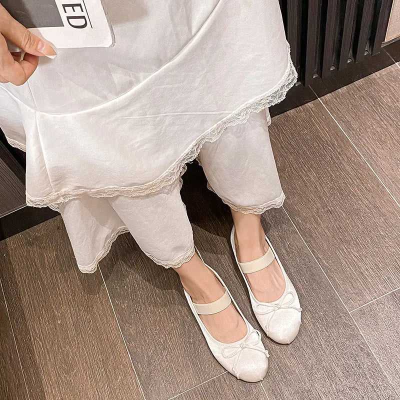 Ivory white satin ballet flats with a delicate bow and elastic instep strap, worn by a person in a relaxed lifestyle setting with draped white fabric.