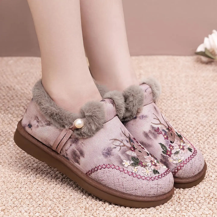 Jevok embroidered plush slippers in a soft pinkish-mauve with intricate floral and deer embroidery, featuring grey faux fur trim and a single pearl accent on a textured rug background.