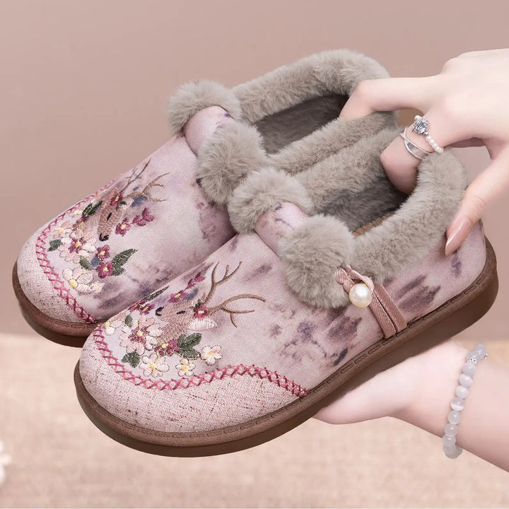 JEVOK embroidered plush slippers in pinkish-mauve with intricate floral and deer motifs, grey faux fur trim, and pearl accent, held by hands showing inner lining.