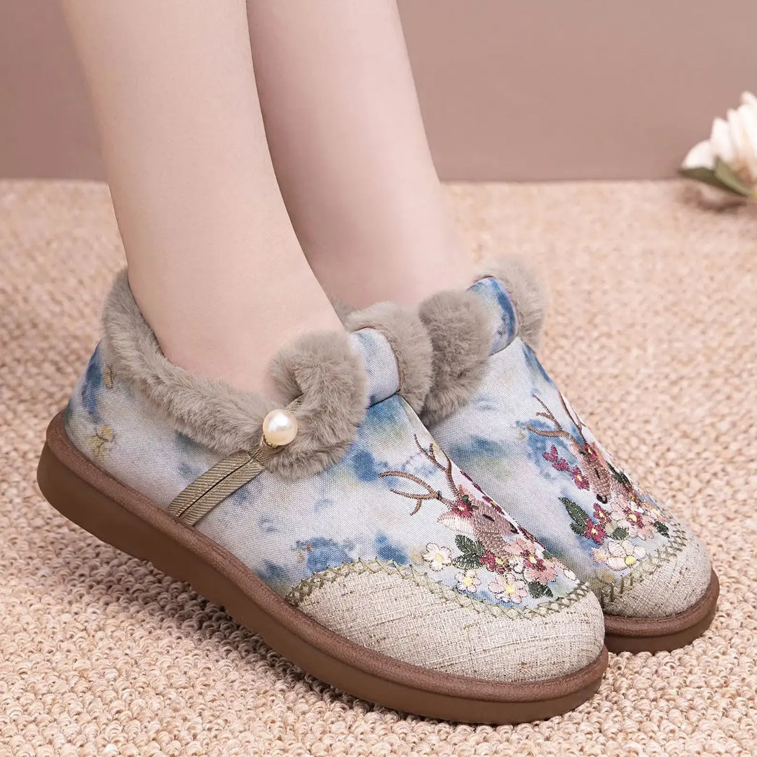 Jevok embroidered plush slippers for women, featuring intricate deer and floral embroidery, soft grey faux fur trim, and a pearl accent, shown on feet with a textured rug background.