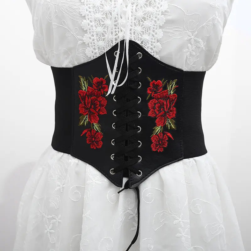 Black corset-style waist belt featuring vibrant red floral embroidery, a central lace-up front, and elasticated sides, displayed on a white dress.