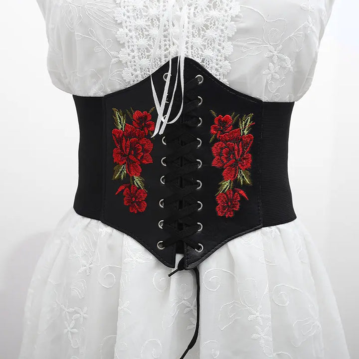 Black corset-style waist belt featuring vibrant red floral embroidery, a central lace-up front, and elasticated sides, displayed on a white dress.