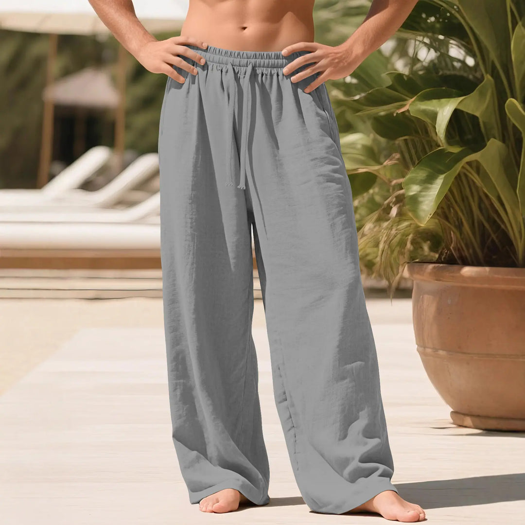 Kaelan men's relaxed fit linen blend trousers in grey, shown on a model in a lifestyle setting with lounge chairs and tropical plants.