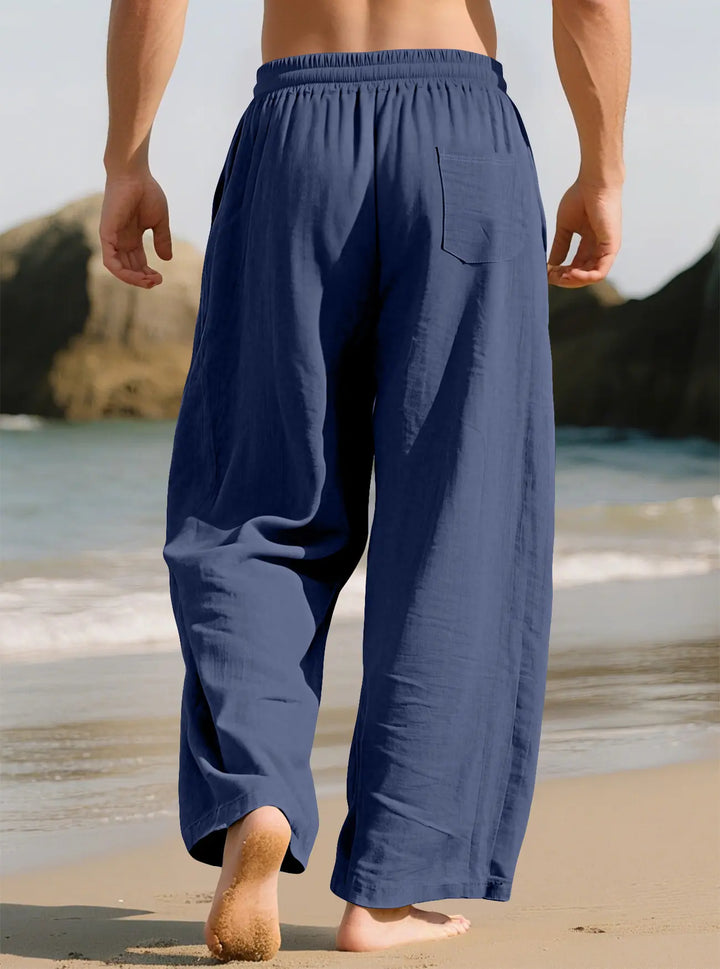 Model from the back wearing navy blue Kaelan linen-blend relaxed-fit trousers on a sandy beach.