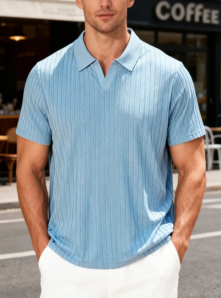 Man wearing a light blue, textured knit V-neck polo shirt with short sleeves and vertical ribbing, styled with white pants in an outdoor urban setting.