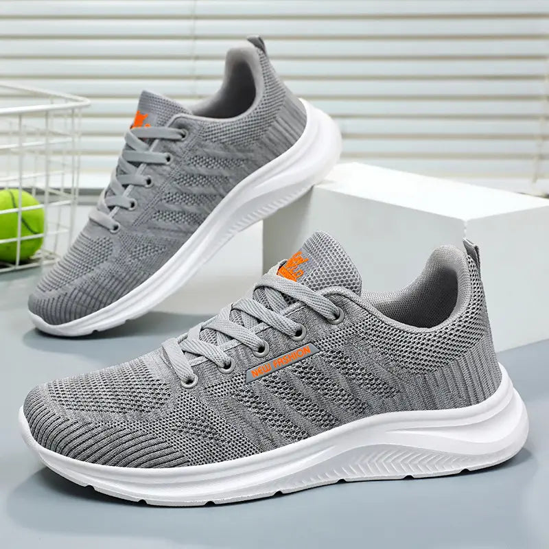 Pair of grey Kaelen breathable mesh trainers with white soles, shown from a three-quarter angle on a studio background. Features tonal laces and subtle orange branding.