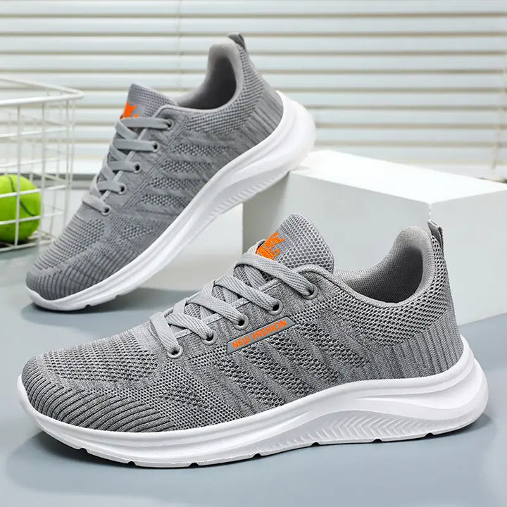 Pair of grey Kaelen breathable mesh trainers with white soles, shown from a three-quarter angle on a studio background. Features tonal laces and subtle orange branding.