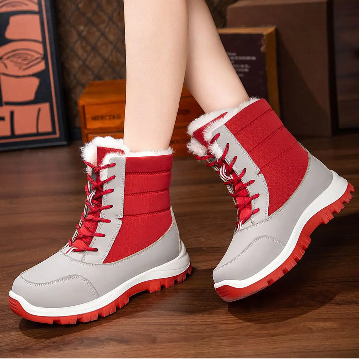 Red and grey mid-calf Kaelen winter snow boots with white faux fur lining and red lace-up closure, worn on feet with legs visible against a warm wooden floor background.