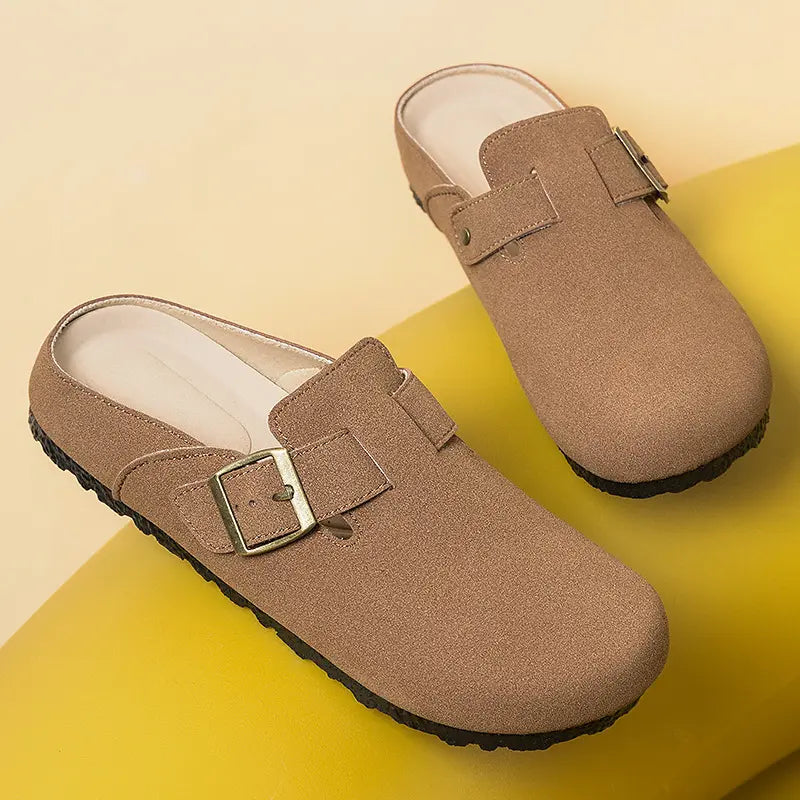 A pair of taupe suede-effect slip-on mules with closed round toes and adjustable antique brass buckle straps, shown from a three-quarter angle on a matte yellow studio background.