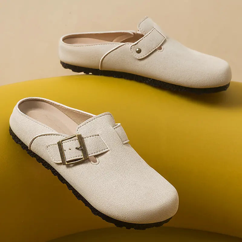 A pair of Kaelen taupe suede-effect slip-on mules with an adjustable antique brass buckle, rounded toe, and flat textured sole, shown from a three-quarter angle on a yellow background.