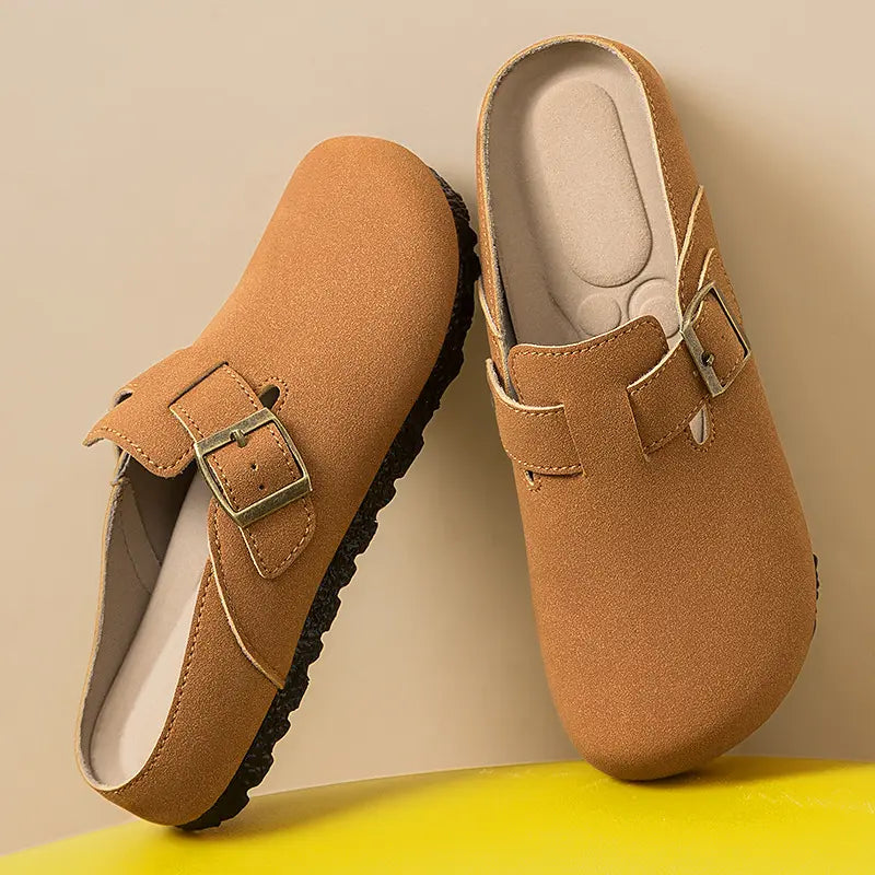 Pair of taupe suede-effect slip-on mules with adjustable antique brass buckle, round toe, and textured black flat sole, shown in a studio setting.