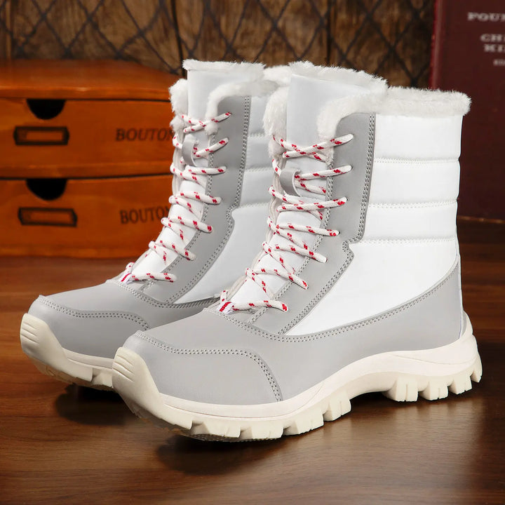Pair of Kaelen mid-calf winter boots in white and light grey, featuring white faux fur trim, red and white patterned lace-up closure, and thick white and red lugged rubber outsoles. These waterproof snow boots are displayed on a wooden surface.