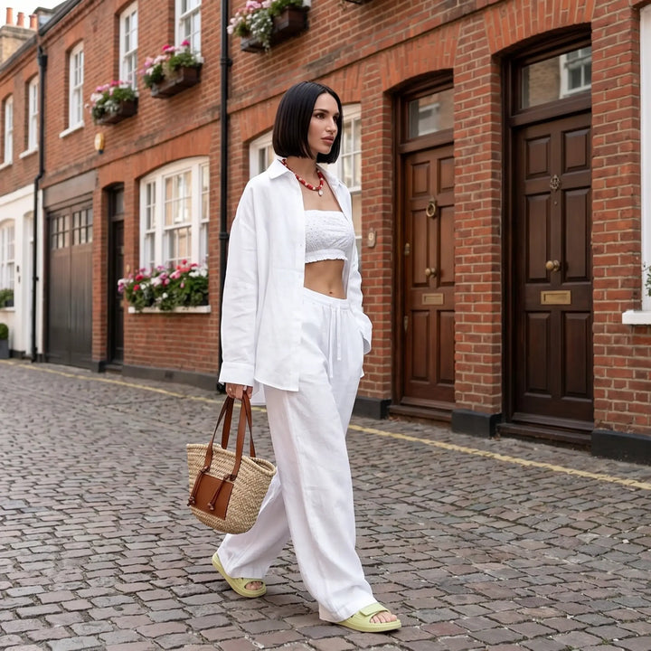 Woman walking on a cobblestone street wearing a white Kaelen linen-cotton relaxed shirt and wide-leg trousers set, holding a straw bag.