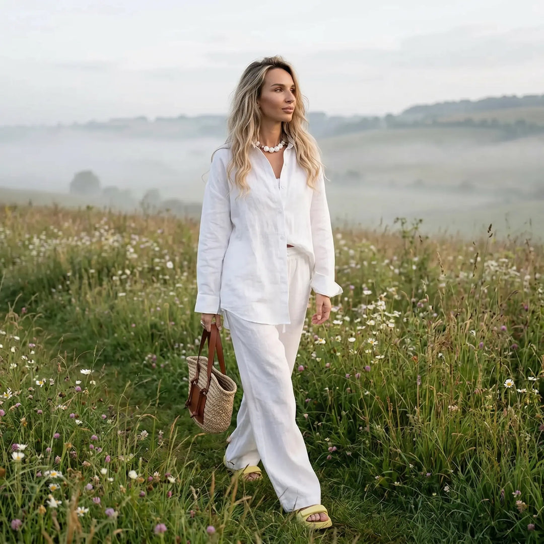 A woman walks through a misty meadow filled with wildflowers, wearing a pure white Kaelen linen-cotton blend relaxed shirt and wide-leg trousers. The outfit embodies effortless style and comfort.
