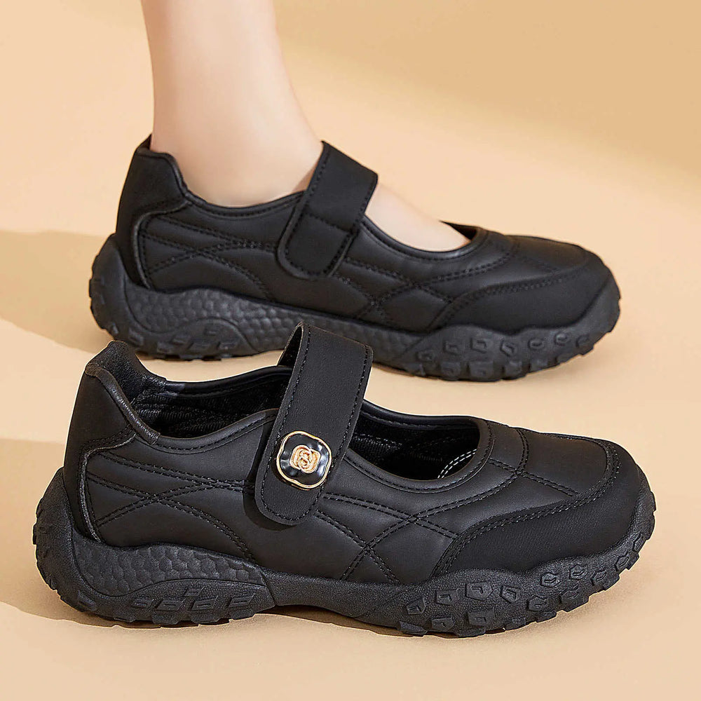Kaelis black quilted Mary Jane flats with adjustable hook-and-loop strap and chunky rubber sole, shown from a three-quarter view with one shoe on a foot in the background.