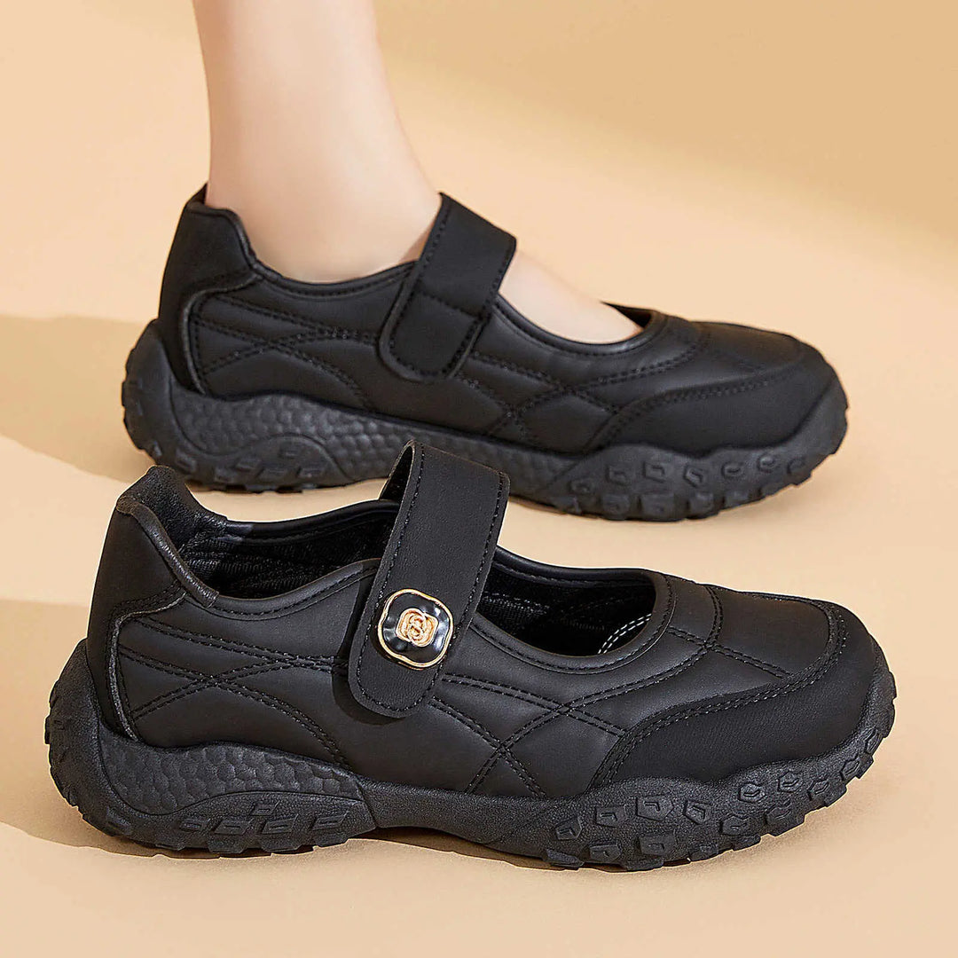 Kaelis black quilted Mary Jane flats with adjustable hook-and-loop strap and chunky rubber sole, shown from a three-quarter view with one shoe on a foot in the background.