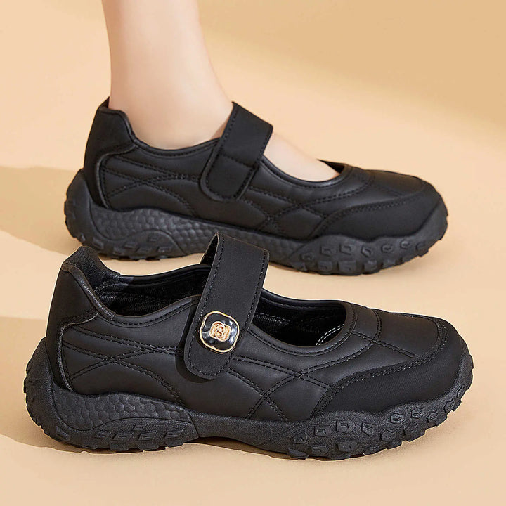 Kaelis black quilted Mary Jane flats with adjustable hook-and-loop strap and chunky rubber sole, shown from a three-quarter view with one shoe on a foot in the background.